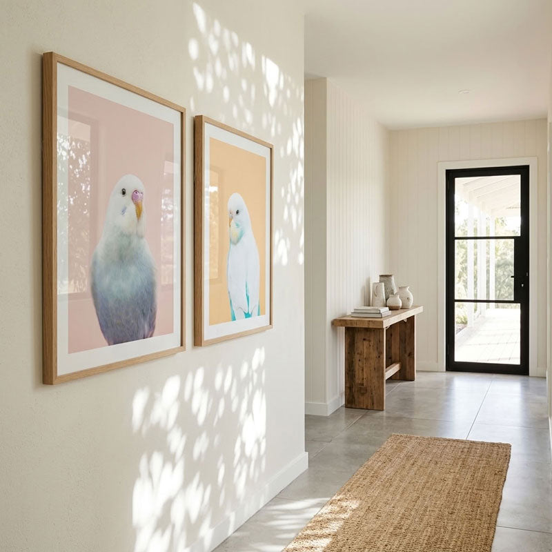 Two framed wall art prints featuring white birds on soft pink and yellow backgrounds, displayed in a modern hallway with natural leaf shadows on the wall.
