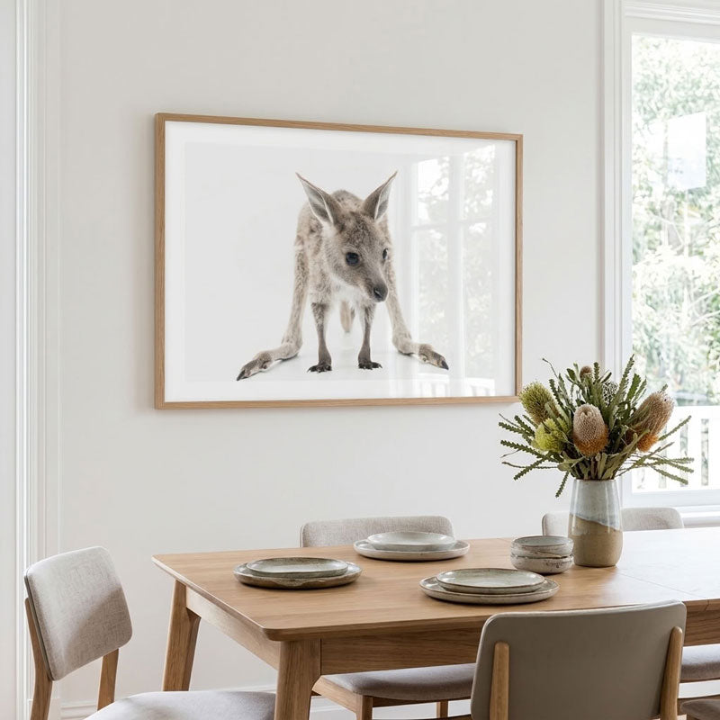 A framed wall art print of a kangaroo hanging above a wooden dining table in a bright, modern room with chairs and a vase of flowers.