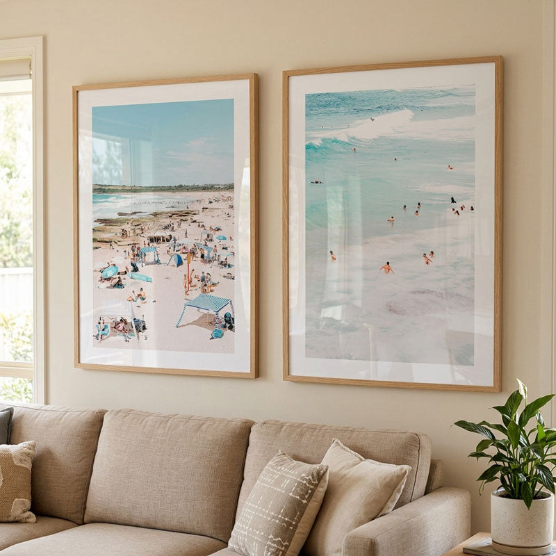 Two framed beach art prints hanging on a beige wall above a sofa in a living room. The left print shows a crowded beach scene with people and umbrellas, and the right print shows a more serene ocean wave scene with small swimmers.