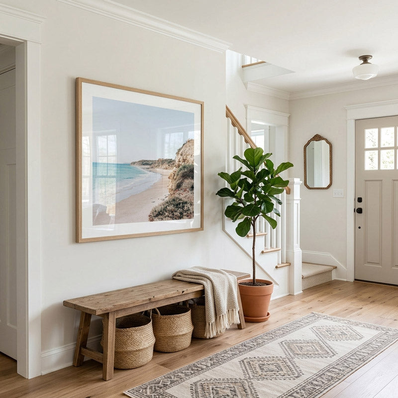 A large framed beach landscape print hangs on a white wall in a bright, modern entryway. Below the frame is a rustic wooden bench with two woven baskets and a draped throw blanket, next to a potted fiddle-leaf fig plant.