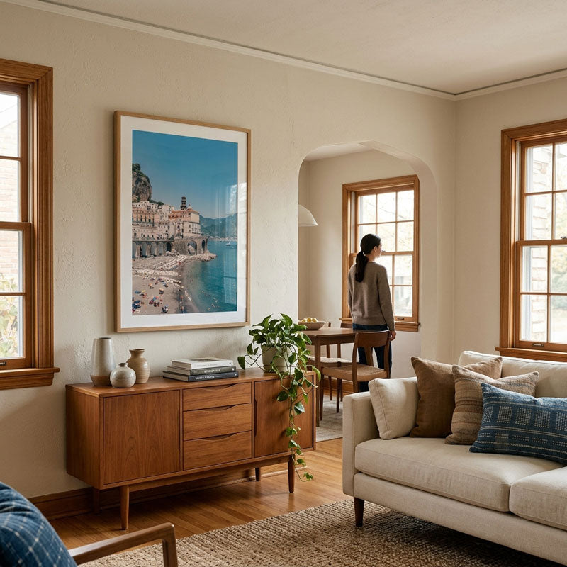 A large framed art print depicting a coastal town scene hangs on a beige wall in a modern, sunlit living room with wood floors, a mid-century sideboard, and a cream sofa.