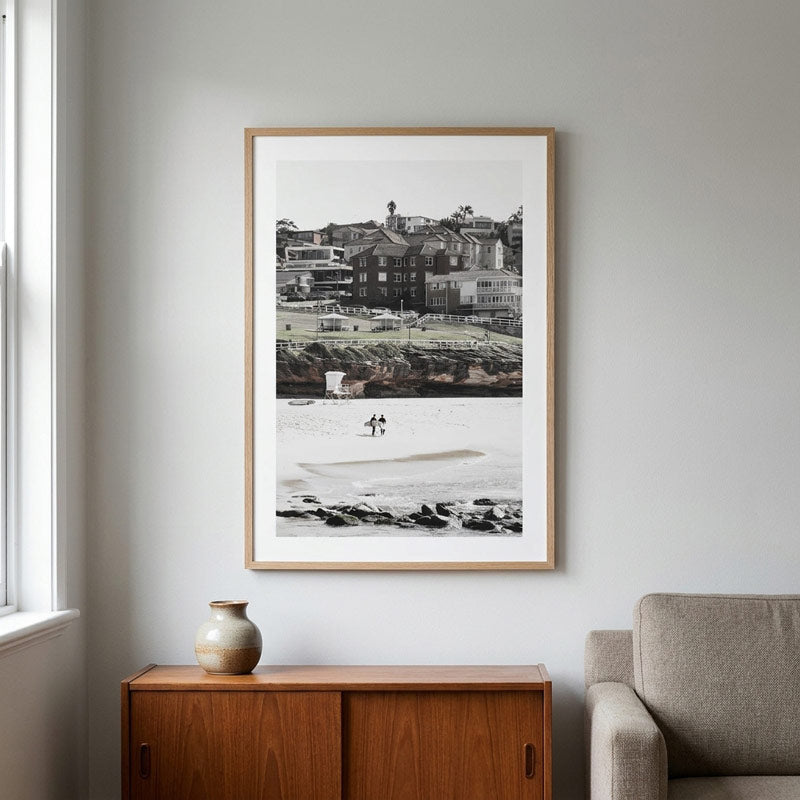 A framed black and white coastal photograph print with a light wood frame, displayed on a light gray wall above a wooden cabinet in a modern living room setting.