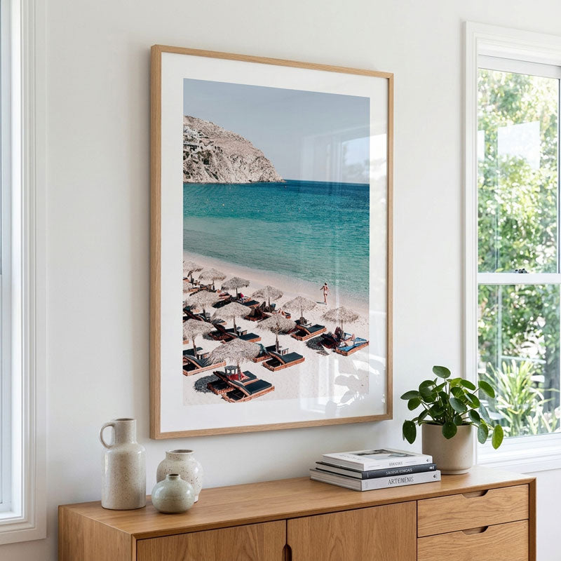 A framed beach art print hangs on a white wall above a wooden console table. The print shows a sunny beach with lounge chairs, umbrellas, turquoise water, and a rocky cliff.