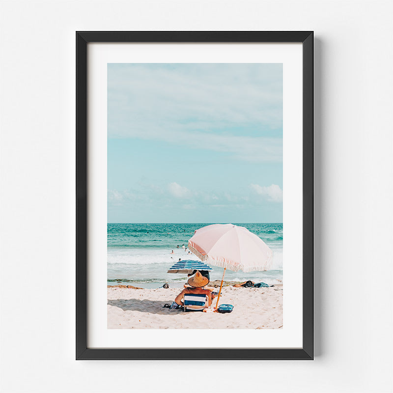 Framed photograph of a person sitting under an umbrella on a beach with ocean view.