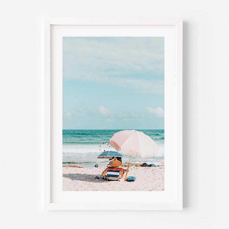 Framed artwork of a beach scene with an umbrella and person sitting under it.