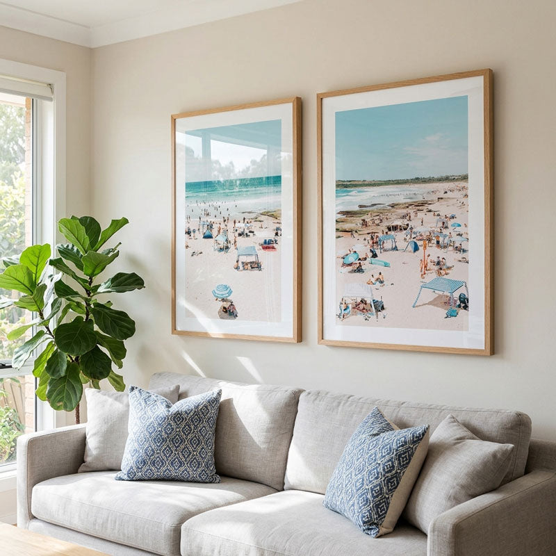 Two framed beach prints hanging on a beige wall above a light gray sofa with blue patterned pillows, next to a large green plant.