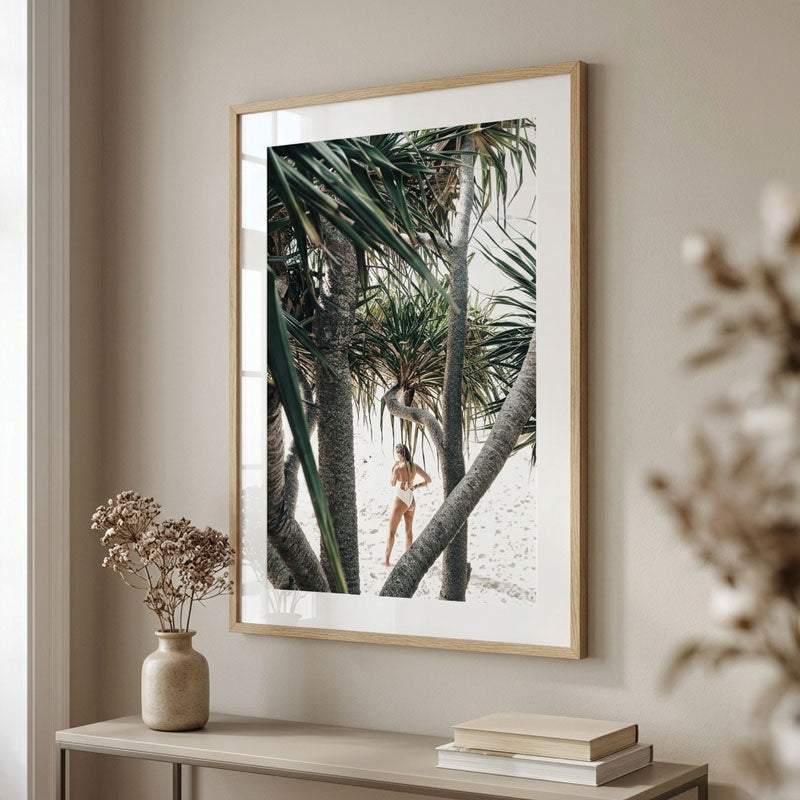 A framed beach photograph featuring a woman standing among palm trees on a sandy shore, displayed on a beige wall above a console table with books and a vase.