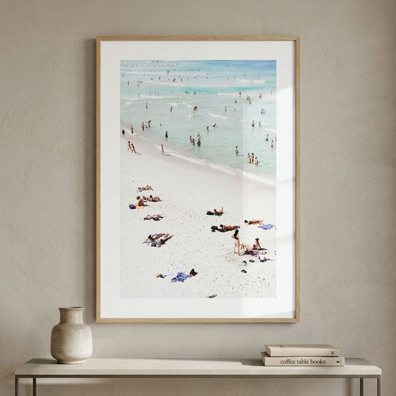 A framed beach print hanging on a textured beige wall above a console table with a vase and books. The artwork shows a sandy beach with small figures swimming and relaxing in the turquoise ocean.