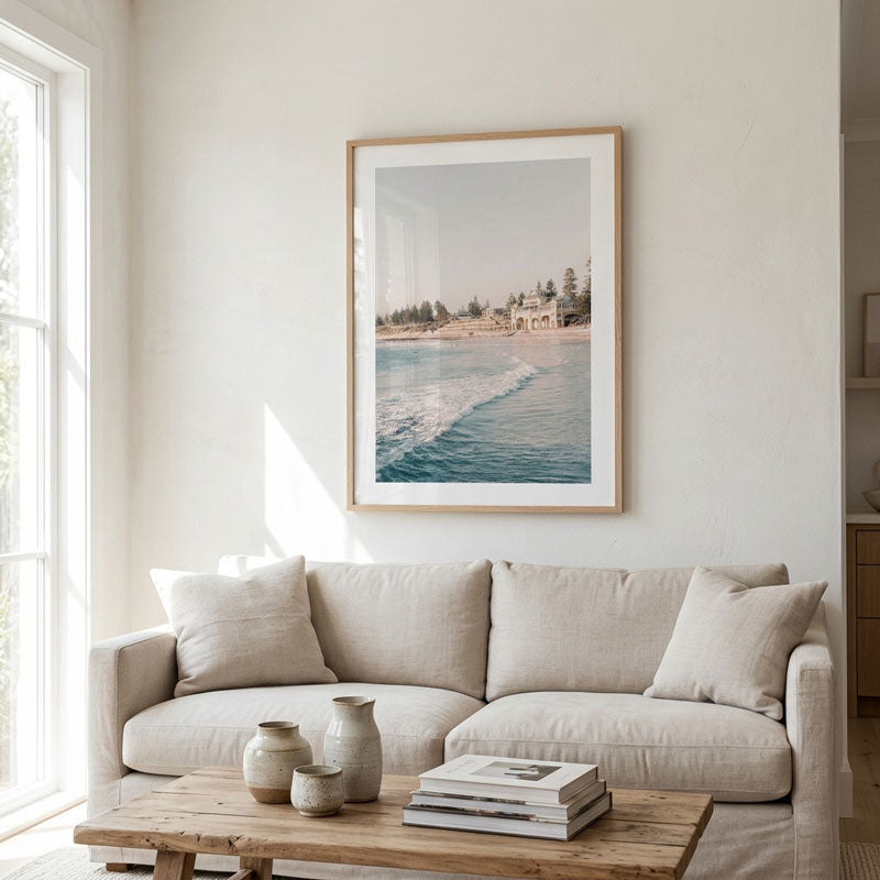 A framed art print depicting a coastal beach scene with blue water and a sandy shore, hanging on a light beige wall above a neutral beige sofa in a bright, modern living room.