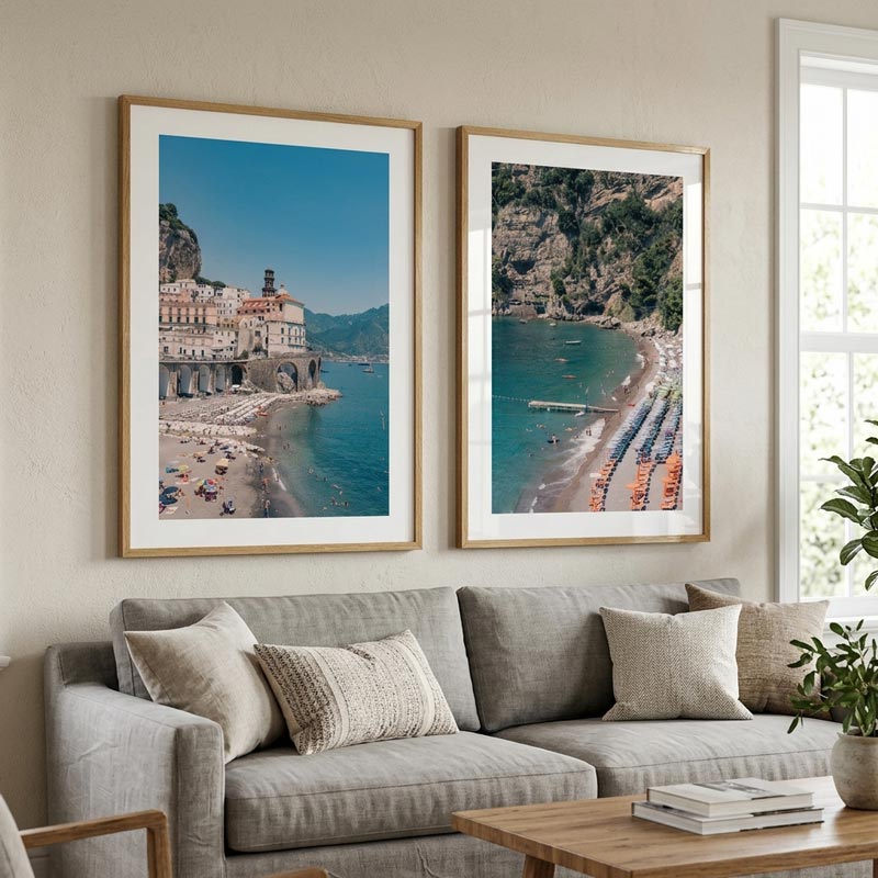 Two framed coastal art prints hang side by side above a light gray sofa in a bright living room. The left print shows a seaside town with a beach and blue water, and the right print shows a rocky cove with a beach and umbrellas.