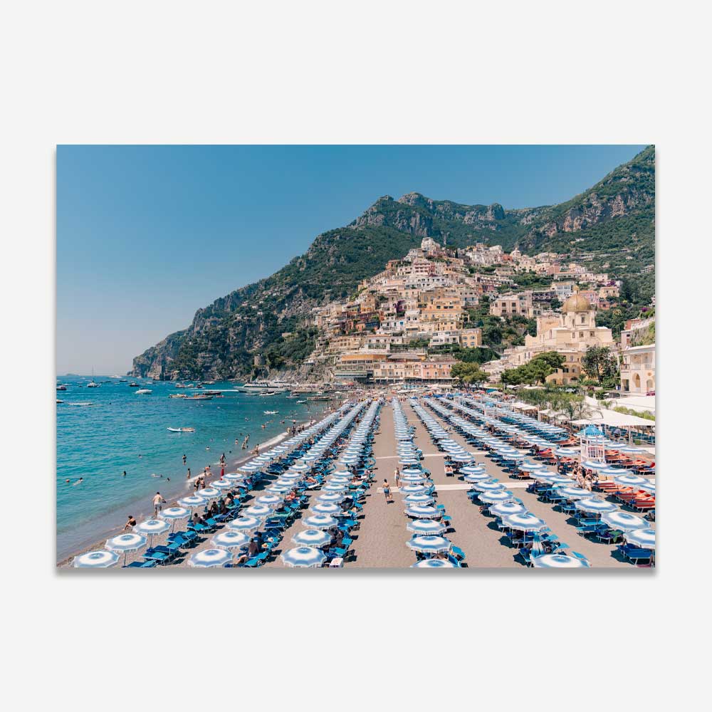 Positano beachfront scene with blue and white beach umbrellas, sunbathers, and crystal-clear waters against the picturesque coastline – capturing the essence of carefree Italian summer days.









