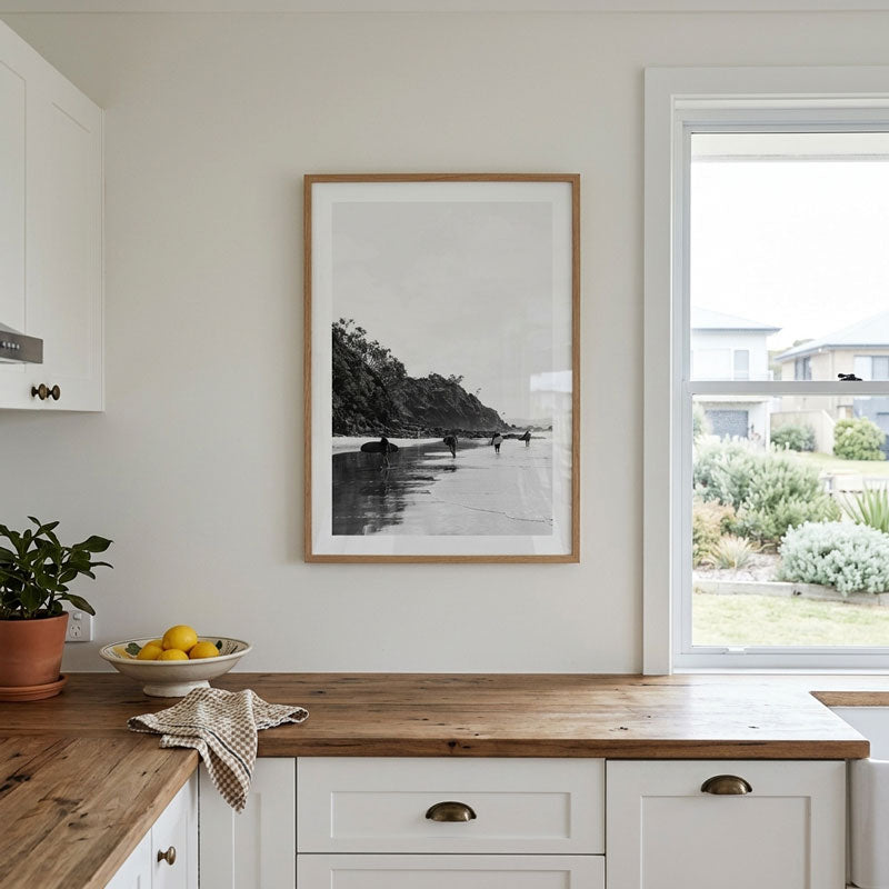 A framed black and white landscape photograph print with a light wood frame hanging on a white kitchen wall above a wooden countertop.