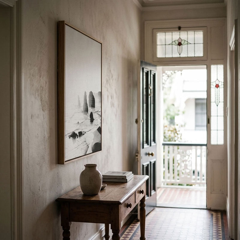 A large vertical black and white framed art print hangs on a textured beige wall in a classic hallway, above a dark wood console table with a ceramic vase and books. The hallway leads to an open door with stained glass and a view of a balcony.