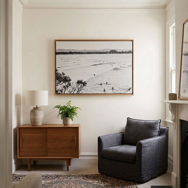 A large black and white framed beach photograph hangs on a cream wall in a styled living room with a wooden sideboard, lamp, potted plant, and a dark gray armchair.