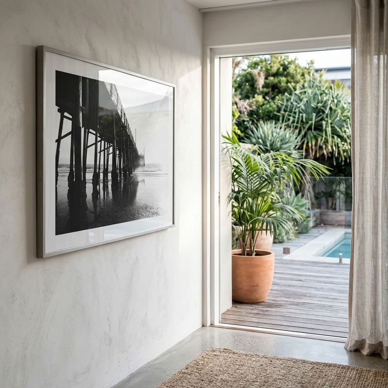 A large framed black and white photograph of a pier structure hanging on a textured light gray wall in a modern interior, with an open doorway revealing a wooden deck, potted plants, and a pool outside.