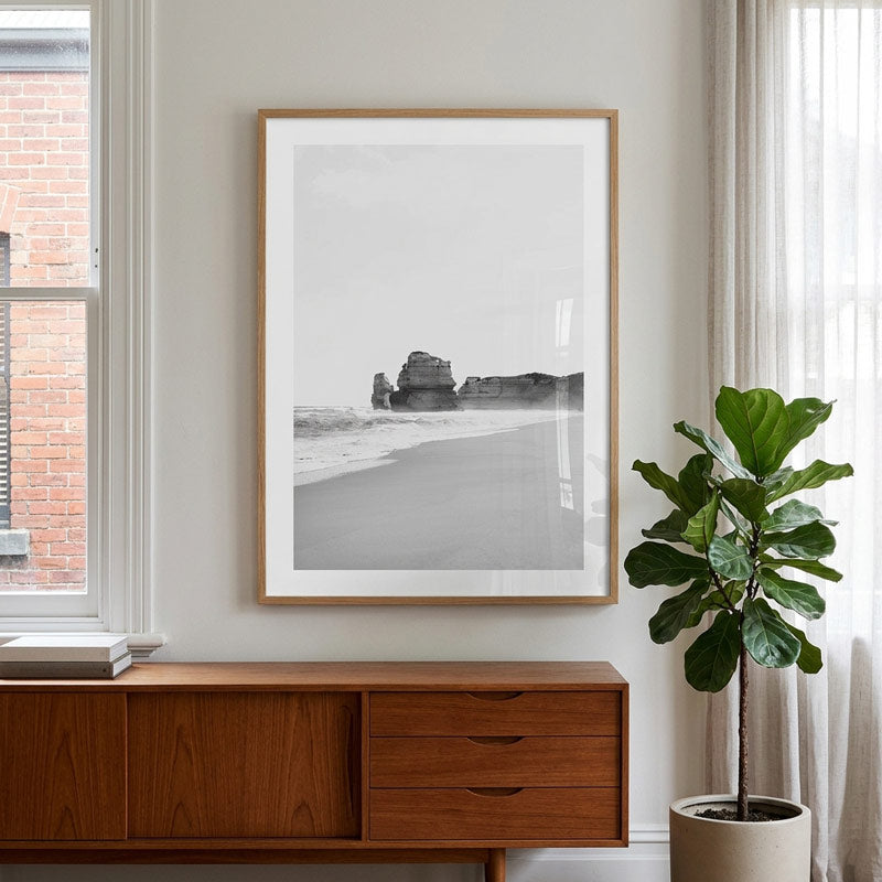 A framed black and white beach photograph with sea stacks hangs on a light wall above a wooden sideboard, with a potted fiddle-leaf fig plant to the right and a window on the left.