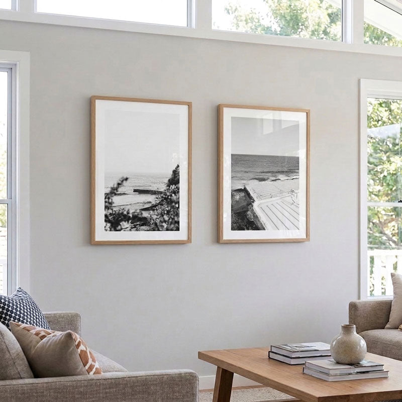 Two framed black and white beach photographs with light wood frames hang on a light gray wall in a modern living room setting.
