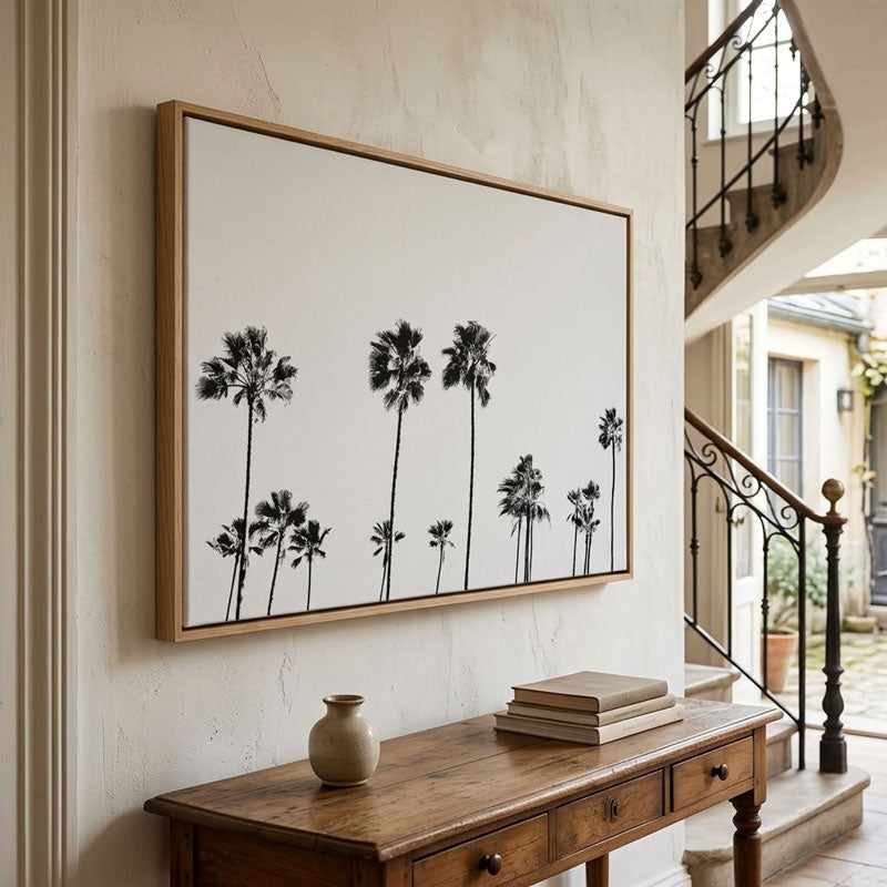 A framed wall art print featuring black palm tree silhouettes on a white background, hung in a warm interior hallway above a wooden console table.