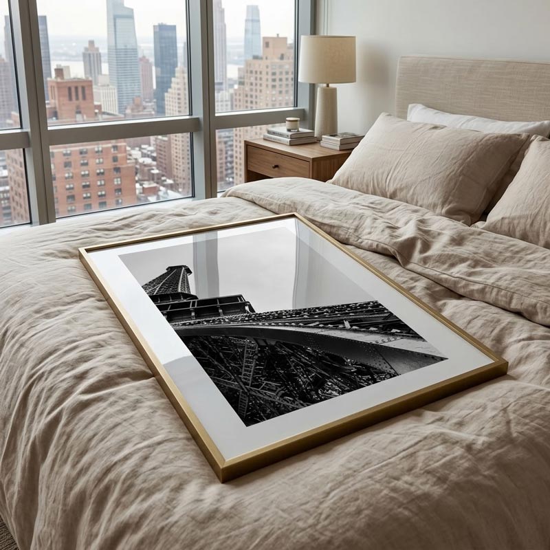 A framed black and white architectural photograph of the Eiffel Tower, set in a gold frame with a white mat, resting on a beige linen bed in a modern bedroom with a city skyline view.