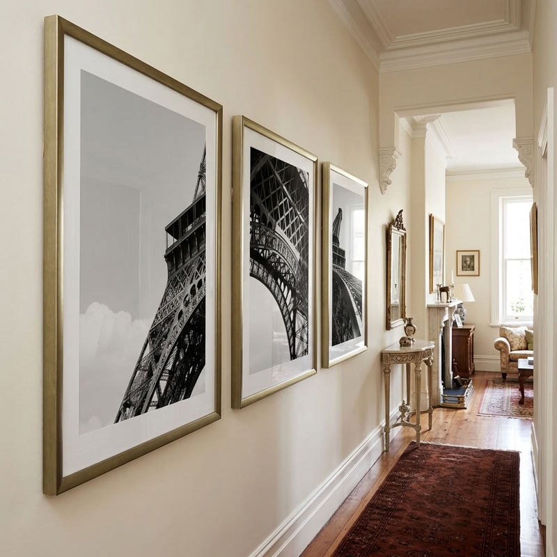 Three large framed black and white architectural photographs of the Eiffel Tower hanging in a hallway with cream walls, wood floors, and a dark rug.