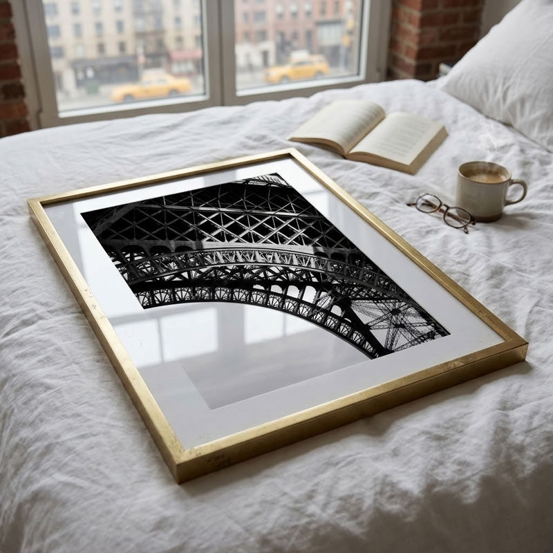 A framed black and white architectural art print of the Eiffel Tower, set in a gold frame on a white bed alongside an open book, a coffee mug, and eyeglasses.