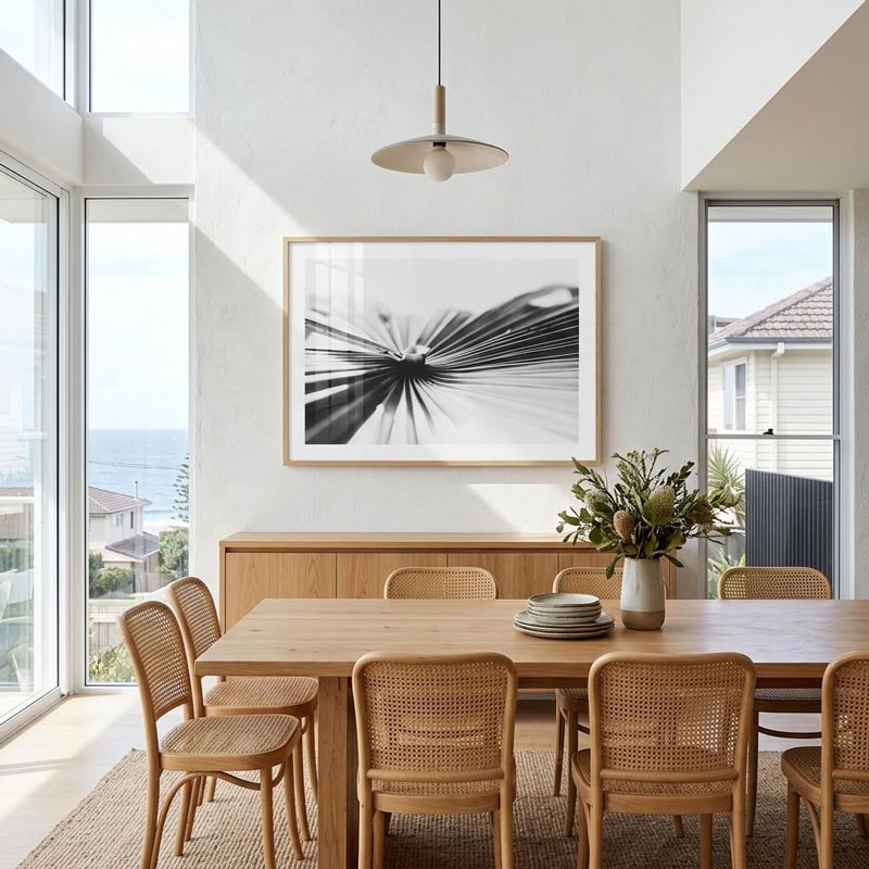A framed black and white abstract art print hangs on a white wall in a bright, modern dining room with a wooden table and woven chairs.