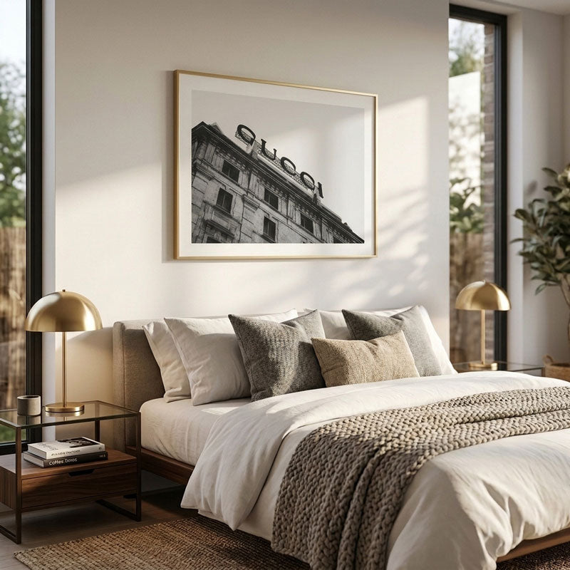 A framed black and white art print of a building with a 'GUCCI' sign, styled in a modern, sunlit bedroom above a bed with neutral-toned bedding.