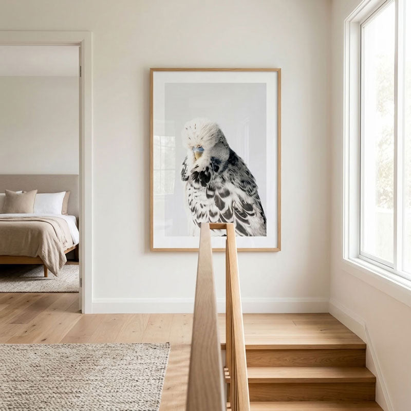 A framed wall art print featuring a black-and-white bird perched on a wooden railing in a bright, modern room with light wood stairs and a window.