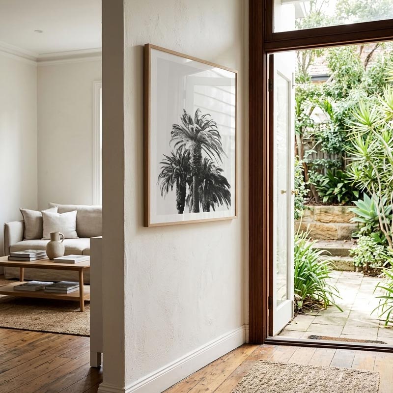 A framed black and white botanical art print of palm trees hangs on a textured white wall in a modern home entryway, with a wooden coffee table and sofa visible on the left and a lush green outdoor patio visible through a glass door on the right.