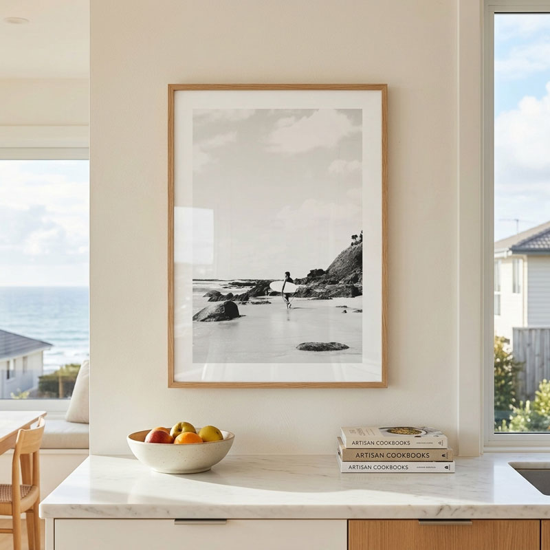 A framed black and white coastal photograph hangs on a cream wall above a white marble kitchen counter with a bowl of fruit and stacked cookbooks. The print has a light natural wood frame and is shown in a bright, modern kitchen setting.