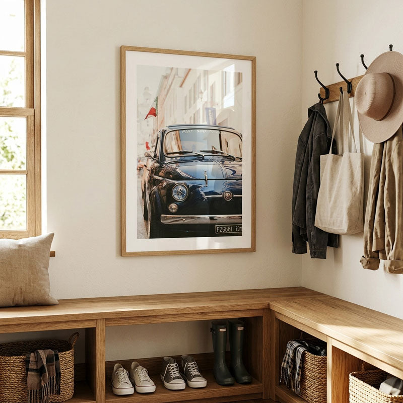 A framed vertical art print of a vintage black car hanging on a beige wall in a styled entryway with wooden benches, baskets, shoes, and hanging jackets and a hat.