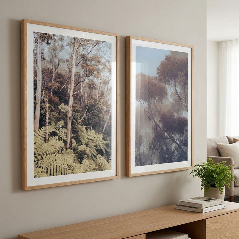 Two framed forest art prints with light oak frames and white mats, displayed side-by-side on a beige wall above a wooden console table in a modern living room setting.