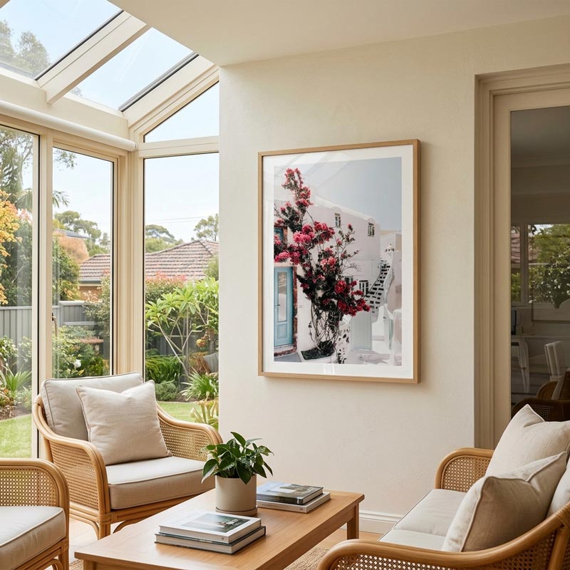 A framed art print depicting red flowers in front of white Mediterranean-style buildings hangs on a beige wall in a bright, modern sunroom with wicker furniture and large skylights.
