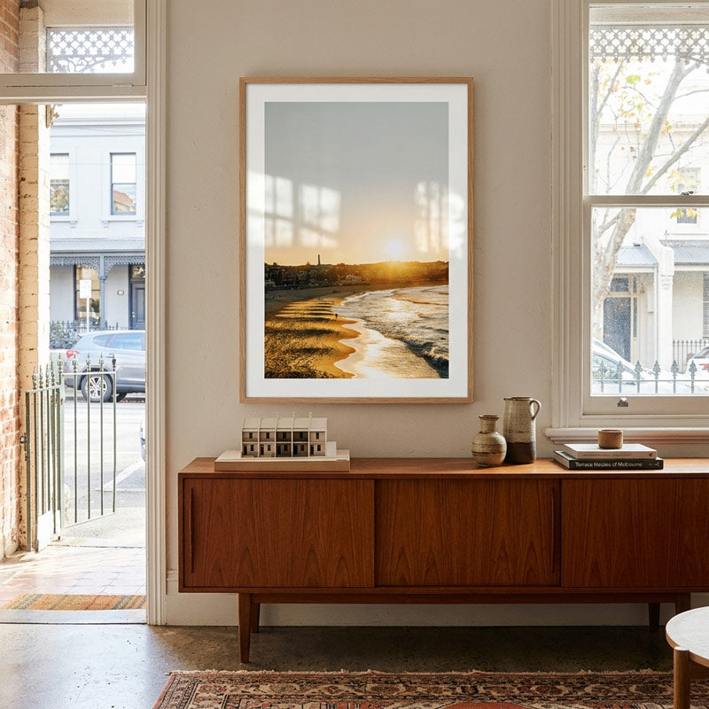 A framed landscape art print featuring a golden sunset over a winding river, displayed on a beige wall above a mid-century modern wooden sideboard in a beautifully styled room.
