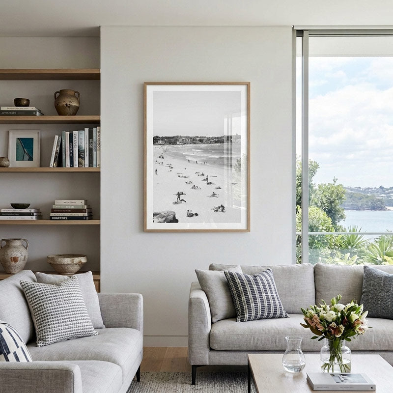 A framed black and white beach print hangs on a light gray wall in a modern living room with two gray sofas, a wooden shelf, and a large window overlooking water.