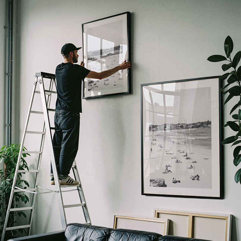 Man on a ladder hanging framed pictures on a wall