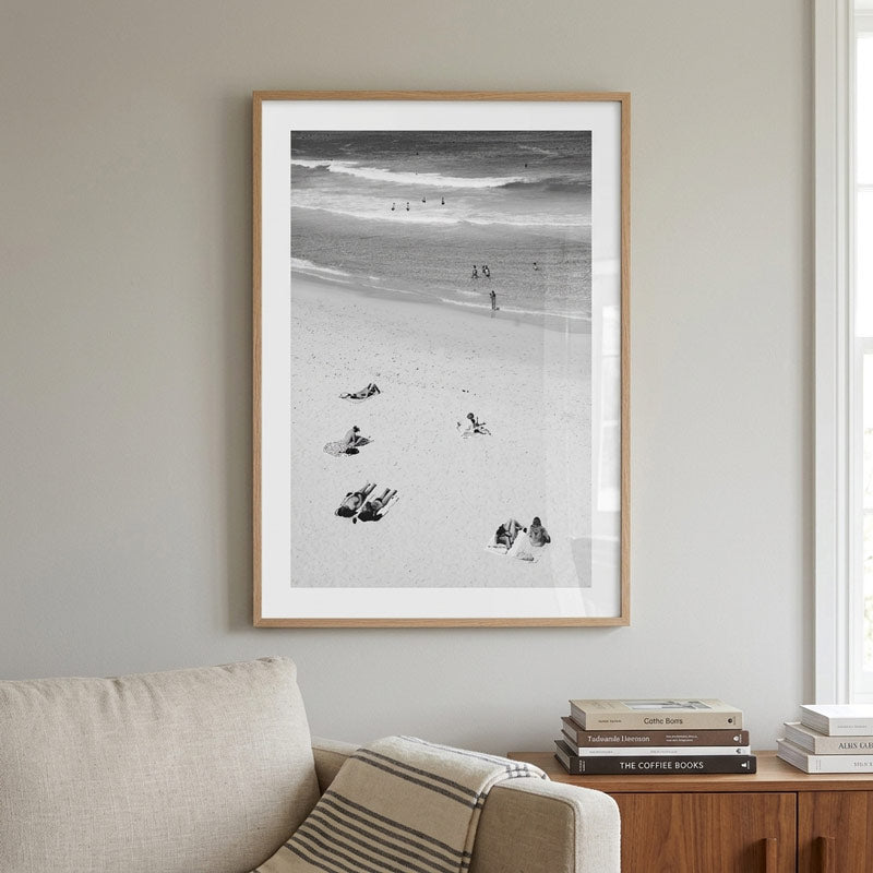 A framed black and white beach photograph hanging on a light gray wall above a beige sofa, with a wooden side table and books below.