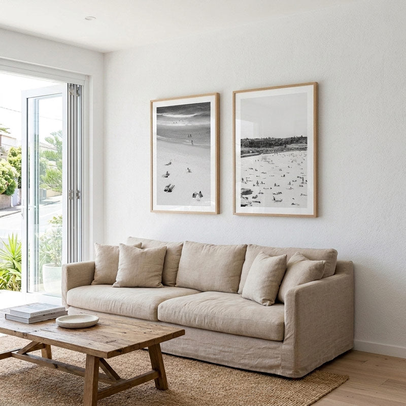 A beige fabric sofa in a bright, modern living room with two framed black and white beach prints on the wall above it, a wooden coffee table, and a window with outdoor views.