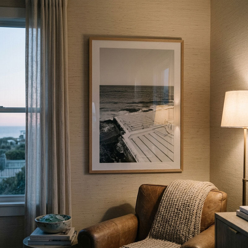 A framed black and white photograph of a seaside pier and rocks, displayed on a beige textured wall in a styled living room with a leather armchair, window, and floor lamp.