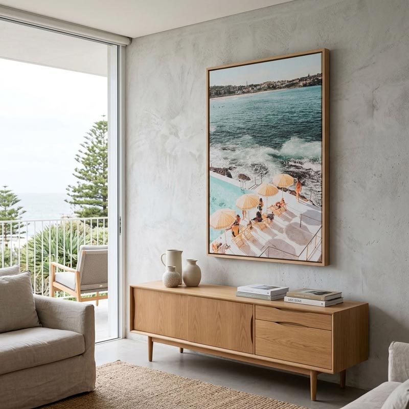 A tall framed coastal art print hangs on a textured light gray wall above a light wood sideboard in a modern living room. The artwork depicts a beachside pool scene with lounge chairs, ocean water, and a shoreline.
