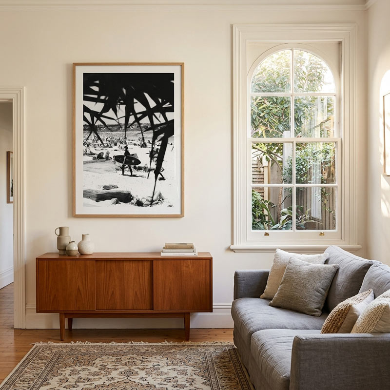 A large black and white framed art print hangs on a cream-colored wall in a beautifully styled living room. The room features a mid-century modern wooden sideboard, a gray fabric sofa with neutral throw pillows, and a patterned area rug on a hardwood floor.