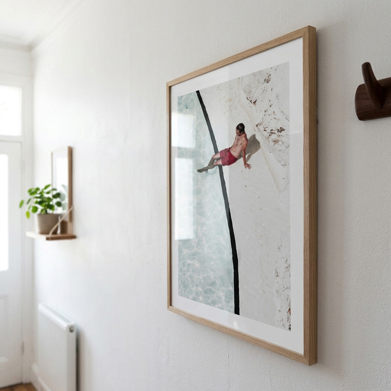 A framed art print featuring a minimalist photograph of a woman in red swimwear on a white and gray background, hung on a white wall in a modern interior setting.