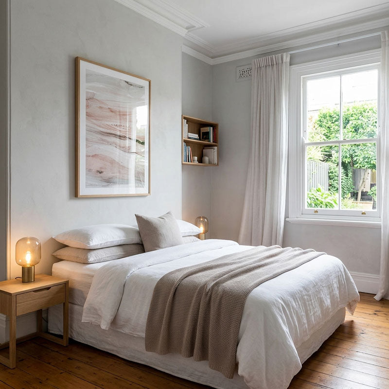 A styled bedroom interior featuring a neatly made bed with white linens and a beige throw blanket, a light wood bedside table with a lit glass lamp, a framed abstract print on the wall, and a window with sheer curtains and visible greenery outside.
