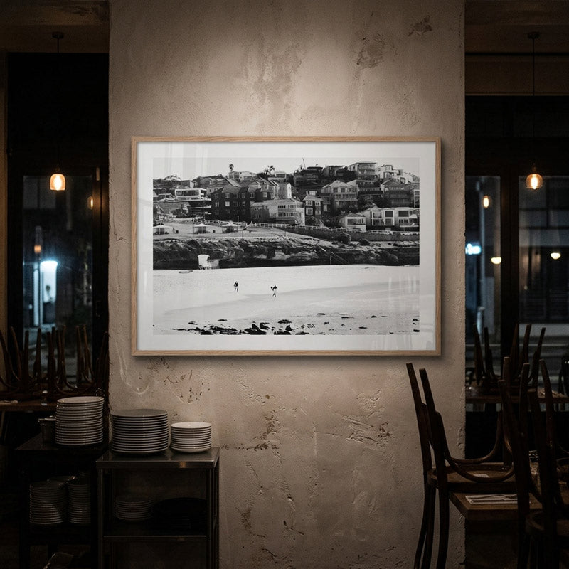 A black and white framed art print depicting a coastal town and beach, displayed on a textured plaster wall in a dimly lit restaurant or cafe setting.