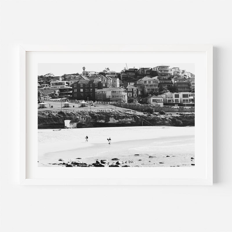 A framed black and white photograph of a coastal town with buildings on a cliffside and a sandy beach in the foreground, shown against a plain white background.