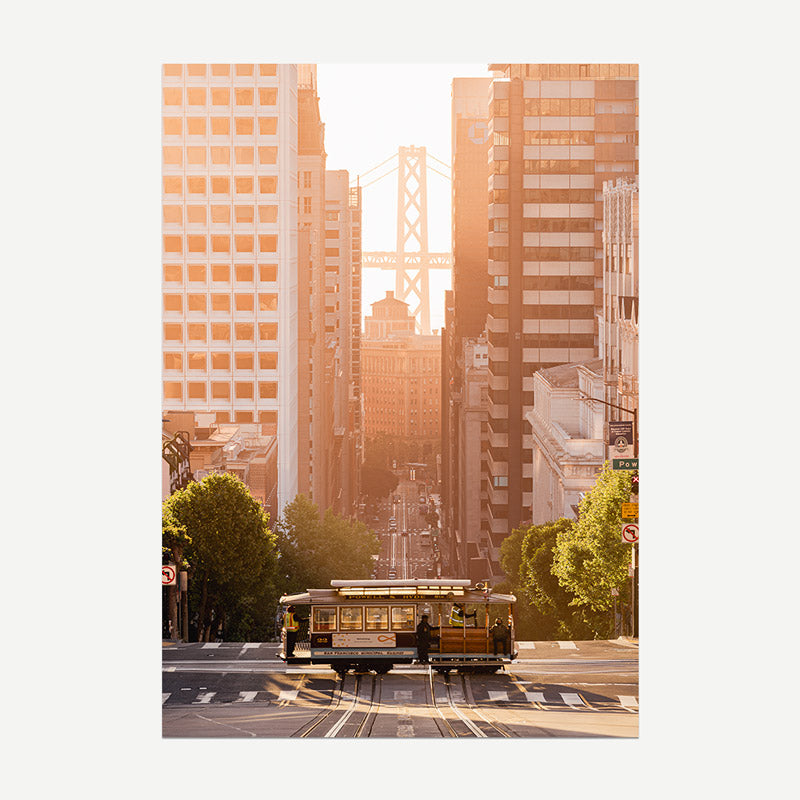 City street with a cable car and tall buildings, including the Bay Bridge, at sunset.