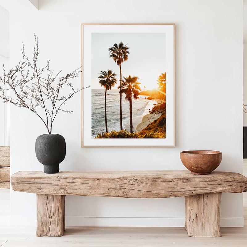 Framed beach scene with palm trees above a wooden bench in a room.