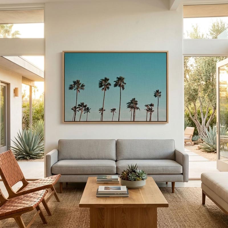 A large framed art print featuring a row of tall palm trees against a turquoise blue sky, hung on a white wall above a light gray sofa in a modern, sunlit living room.