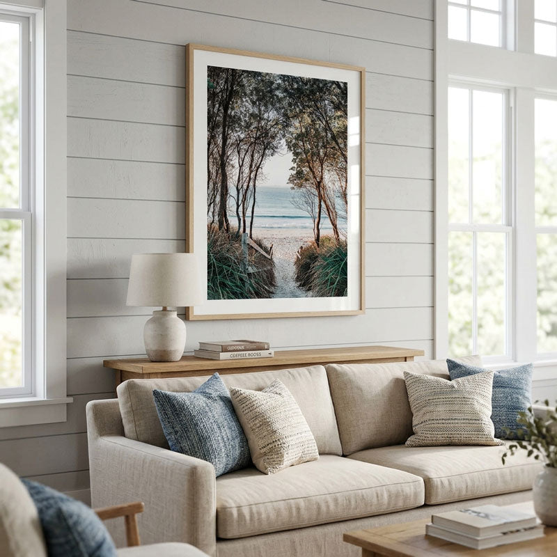 A framed vertical art print depicting a forest dune path leading to the ocean, styled on a white shiplap wall above a beige sofa in a bright, modern living room.