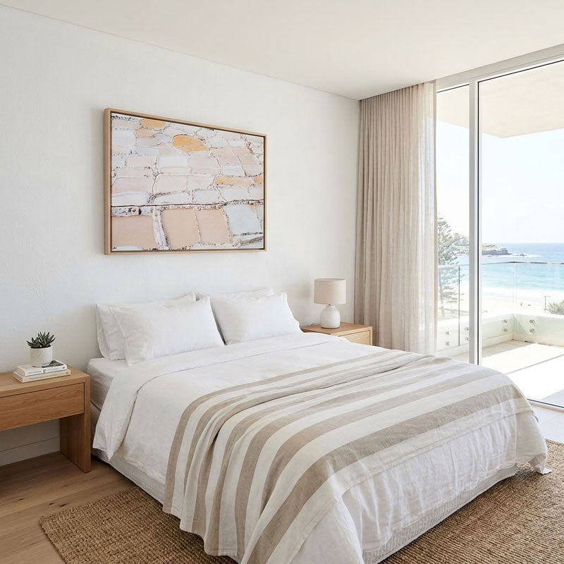 A modern bedroom featuring a large framed abstract art print with beige, white, and gold tones hanging above a neatly made bed with white linens and a beige striped throw. The room has light wood flooring, matching wooden nightstands, a white table lamp, and a large sliding glass door looking out to the ocean.